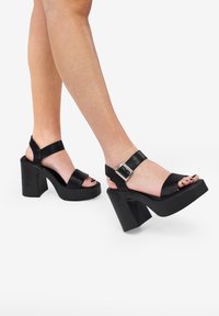 Sandalias de tacón alto con plataforma negra, con correas anchas, hebilla ajustable, diseño de punta abierta y gruesos tacones cuadrados. Textura de cuero suave.