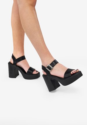 Sandalias de tacón alto con plataforma negra, con correas anchas, hebilla ajustable, diseño de punta abierta y gruesos tacones cuadrados. Textura de cuero suave.