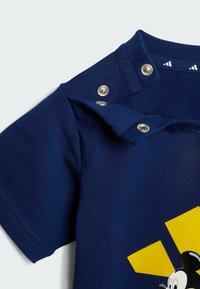 Mörkblå bomullströja med en grafik av Mickey Mouse och en gul geometrisk design. Knäppning på axeln. Slen yta.