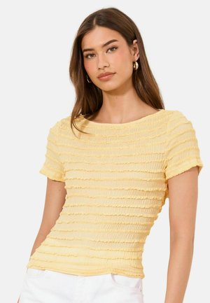 Young woman with long brown hair wearing gold hoop earrings and a short-sleeve, textured yellow top, posing against a plain background.