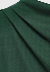 Tejido verde oscuro con textura visible, doblado diagonalmente sobre un fondo claro liso.