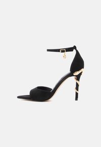 Zwarte suède sandaal met hoge hak, puntige neus, enkelbandje en gouden accenten op de hak. Soepele textuur, elegant ontwerp.