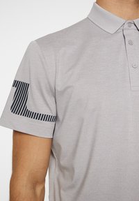 Gray polo shirt with a textured fabric, featuring a black striped design on the left sleeve and a three-button placket.