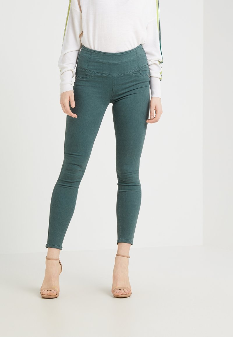 Tealowe legginsy z wysokim stanem, o gładkiej fakturze, z przeszyciami oraz zwężanym krojem nogawek, w połączeniu z beżowymi sandałami na obcasie.