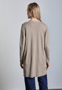 Langer beiger Pullover mit lockerer Passform, gerippten Bündchen und geradem Saum, kombiniert mit dunkelblauen Jeans. Der Stoff wirkt weich und texturiert.