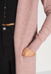 Lange roze cardigan met geribbelde manchetten en een voorkwak. Het materiaal lijkt zacht met een soepele textuur tegen een zwarte top en broek.