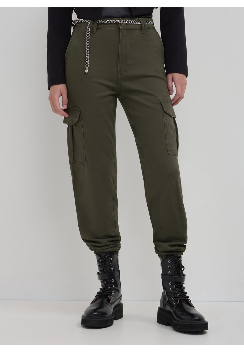 Pantalon cargo vert olive avec poches à rabat, associé à des bottes de combat noires à lacets et une ceinture chaîne argentée.