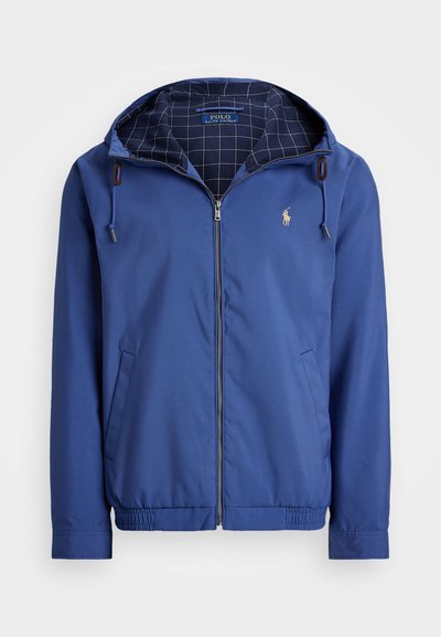 Blå nylonjakke med hette, glidelås foran, elastiske mansjetter og hem. Innvendig har den et rutemønster. Liten logo på brystet.