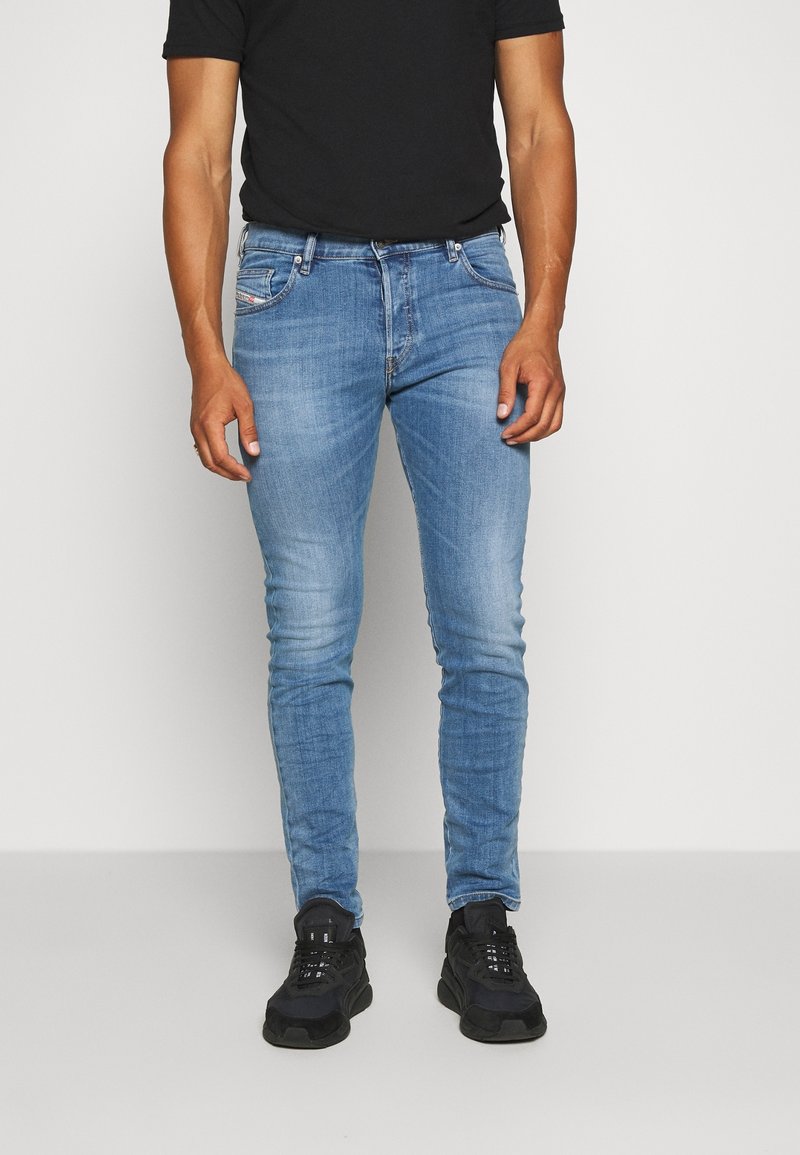 Homme portant un jean slim bleu clair, un T-shirt noir et des baskets noires, debout devant un fond uni.