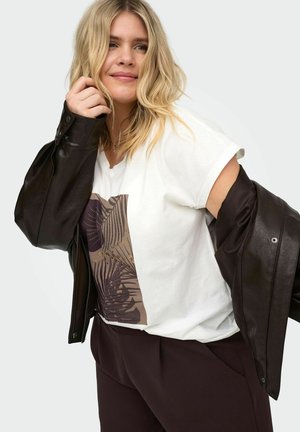 Blondynka w białej koszulce z nadrukiem i ciemnych spodniach wkłada ciemnobrązową skórzaną kurtkę, lekko się uśmiechając.