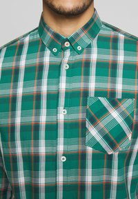 Grøn ternet skjorte med knappet krave; har hvide, grå og røde accenter; inkluderer en lomme med diagonalt ternet mønster.