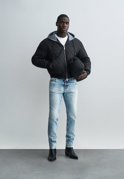 Homme portant une veste matelassée noire avec une capuche grise, une chemise blanche, un jean bleu clair et des bottines noires, tenant un sac noir.