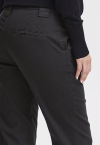 Črne bombažne hlače z gladko teksturo, z zadnjim žepom in topstičenimi šivi, oblečene z temno jopo.