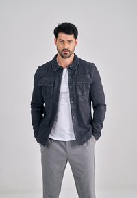 Tmavě modrá džínová bunda s límečkem, předními kapsami a zapínáním na knoflíky, nošená přes bílé tričko a šedé kalhoty.