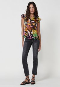 Blusa a stampa floreale con maniche a volant in nero, caratterizzata da vivaci motivi arancioni, rosa e verdi, abbinata a jeans skinny neri e sandali con cinturino.