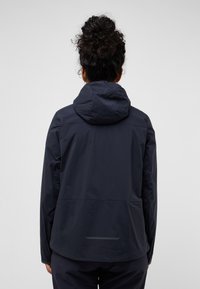 Giacca impermeabile blu navy con cappuccio, polsini elasticizzati e una texture leggera; presenta una striscia riflettente nella parte bassa della schiena.