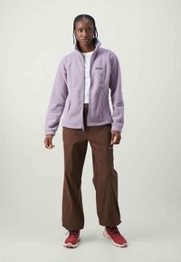 Veste en polaire lilas avec fermeture éclair à l'avant, portée sur un haut blanc, assortie à un pantalon marron et des chaussures rouges. Design simple et fonctionnel.