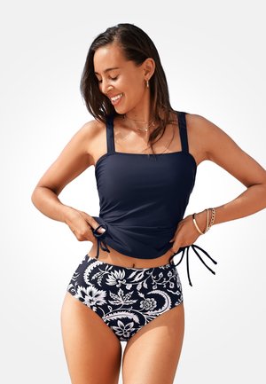 Usměvavá žena si upravuje námořnicky modrý tankini top se zavazováním na bocích, má na sobě černobílý květinový spodní díl bikin s vysokým pasem.