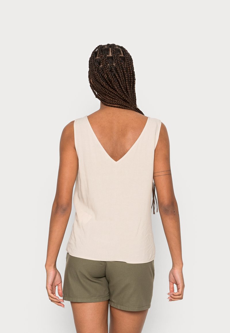 Femme aux cheveux tressés portant un haut beige sans manches avec un dos en V profond et un short vert olive, debout devant un fond uni.