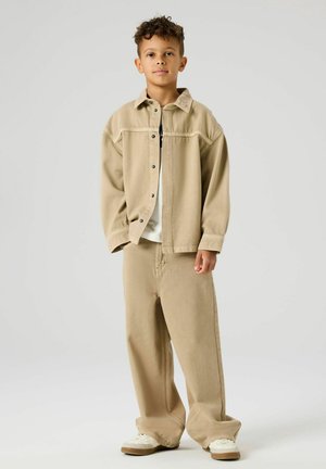 Noor poiss lühikeste lokkis juustega, kandes üle suuruses beežikat jopet ja sobivaid laia säärega pükse, seisab tavalise tausta ees.