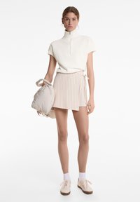 Vitt zip-up-top med korta ärmar, kombinerat med en beige randig minikjol. Mjuka texturer. Modellen håller en ljusfärgad ryggsäck.