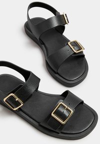 Mustat sandaalit, joissa on teksturoitu tekonahkahihna, kaksi kultaiselta näyttävää solkea, litteat pohjat, avonaiset takapuolet ja minimalistinen muotoilu.