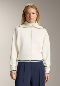 Femeie blondă îmbrăcată cu un pulover crem cu fermoar și guler larg, purtând pantaloni bleumarin, stând pe un fundal neutru.