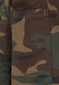 Kámos anyag barna, zöld és fekete árnyalataiban. Bal oldalon négyzet alakú zseb található megerősített varrással.