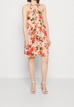 Vrouw draagt een mouwloze roze jurk tot op de knie met een kleurrijk bloemenpatroon en witte open-toe hoge hakken, staand.