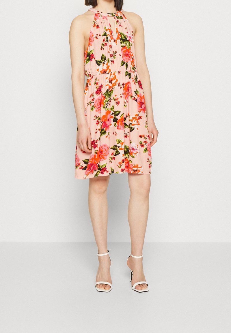 Femme portant une robe rose sans manches, longueur genou, avec un motif floral coloré et des sandales blanches à talons hauts et bouts ouverts, debout.
