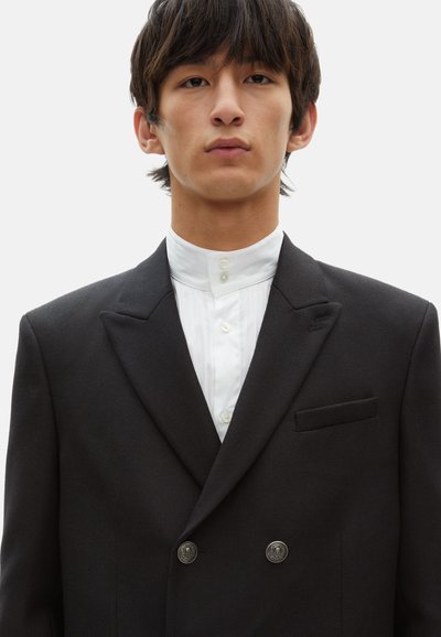 Schwarzes zweireihiges Blazer mit strukturiertem Stoff, mit Reverskragen und silbernen Knöpfen, über einem weißen plissierten Hemd.