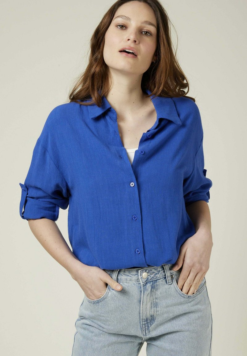 Chemise bleu à boutons avec un tissu texturé, manches retroussées et coupe décontractée, assortie à un jean en denim clair.