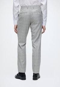 Personne portant un pantalon de costume gris à carreaux, une chemise blanche et des chaussures habillées noires, vue de dos sur un fond uni.