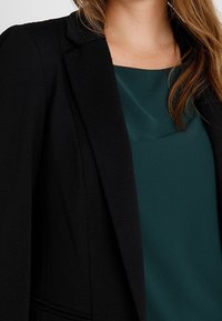 Blazer nero con tessuto texturizzato e rever affilati, indossato sopra un top liscio di colore teal. Concentrati sul design strutturato del blazer e sulle linee pulite.