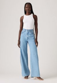 Woman with long braids wearing a white sleeveless top, high-waisted wide-leg light blue jeans, and beige heeled sandals, standing straight.