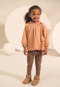 Blouse couleur pêche avec motif floral et col à volants, leggings marron avec ourlets effilochés, et bottes beige hauteur cheville.
