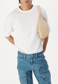 Bolso cruzado beige con un diseño redondeado y detalle de cremallera, usado sobre una camiseta blanca, combinado con pantalones cargo de denim azul.