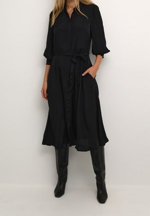 Femme portant une robe midi noire boutonnée avec des manches bouffantes et une ceinture à la taille, associée à des bottes noires montantes.