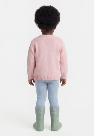 Kind mit lockigem Haar, das einen pinken Pullover, hellblaue Hosen und grüne Gummistiefel trägt, steht mit dem Rücken zum Betrachter auf einem einfarbigen Hintergrund.