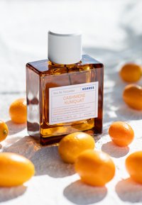 Klar glasflaska med vit kork, etiketterad "Cashmere Kumquat." Omgiven av små kumquats på en texturerad yta. 50 ml storlek.