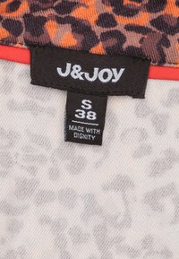Label van een kledingstuk met dierenprint in tinten oranje en bruin, op een beige achtergrond. Het label vermeldt "S 38" en "MADE WITH DIGNITY."