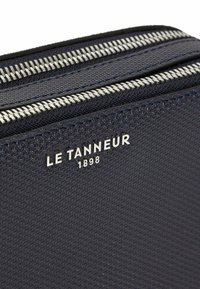 Busta in pelle strutturata blu navy con doppie cerniere argentate e un logo lucido "LE TANNEUR 1898" chiaramente visibile sul fronte.