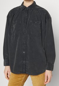 Persona che indossa una camicia button-up in corduroy nera sfumata e morbida, con due tasche sul petto, abbinata a pantaloni in corduroy giallo senape.