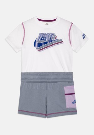 Conjunto de Nike que incluye una camiseta blanca con un logo azul y púrpura, combinada con unos pantalones cortos grises que tienen un bolsillo púrpura y una cinturilla elástica.