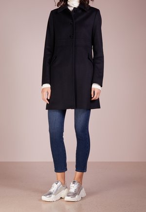 Kvinde iført en sort knælange frakke, hvid rullekrave, blå korte jeans og chunky sølvmetalliske sneakers på ensfarvet baggrund.