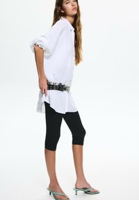 Camicia bianca con maniche arrotolate, leggings neri e infradito verdi. Cintura nera distintiva con dettagli argentati e fori.
