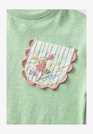 Svijetlozeleni majica s bijelim i zelenim prugastim džepom s ružičastim valovitim rubovima, ukrašena šarenim cvjetnim vezom.