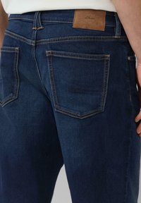 Jean en denim bleu foncé avec une texture lisse, doté d'une patch en cuir étiqueté "s.Oliver", et de poches arrière visibles avec des coutures orange.
