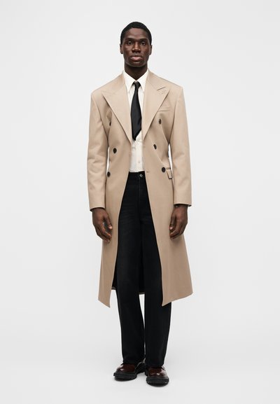 Abrigo cruzado color beige con solapas extragrandes, llevado sobre una camisa blanca y corbata negra. Combinado con vaqueros oscuros y zapatos marrones.