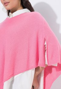 Růžové pletené pončo s bočními rozparky, s texturovaným povrchem a širokým výstřihem do V, vrstvené přes bílou košili.